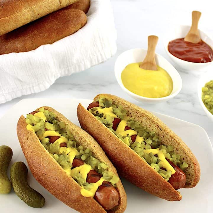 2 Keto Hot Dog Buns with hot dogs, mustard and relish with 2 pickles on side