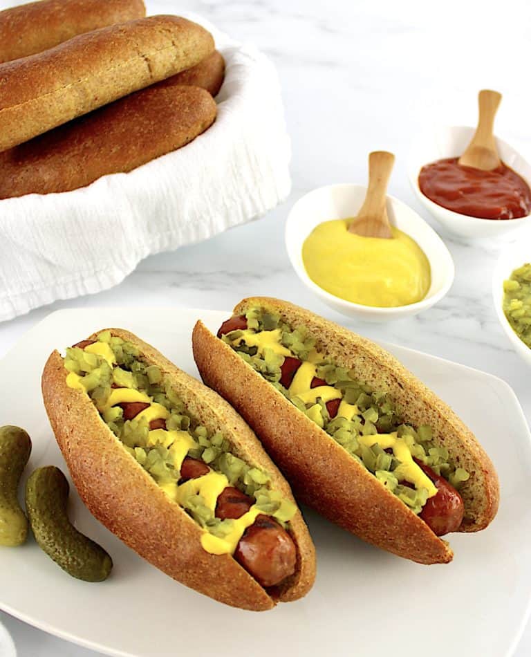 2 Keto Hot Dog Buns with hot dogs, mustard and relish with 2 pickles on side