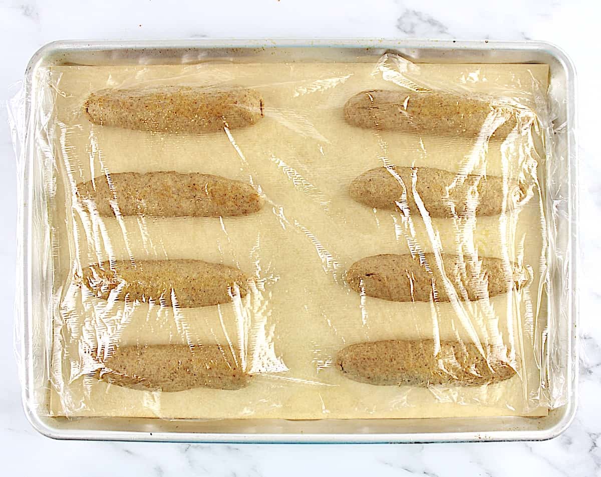 8 hot dog buns dough lined up on baking sheet with plastic wrap on top