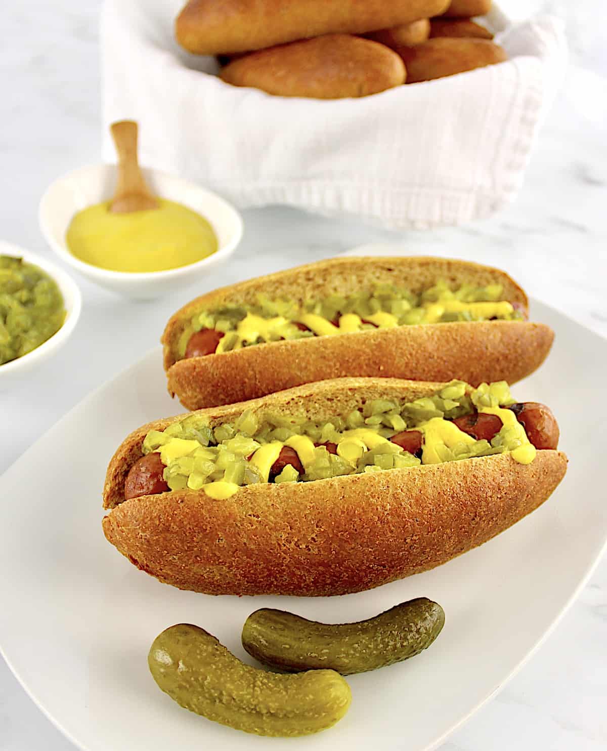 2 Keto Hot Dog Buns with hot dogs, mustard and relish with 2 pickles on side