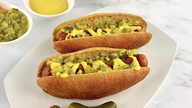 2 Keto Hot Dog Buns with hot dogs, mustard and relish with 2 pickles on side