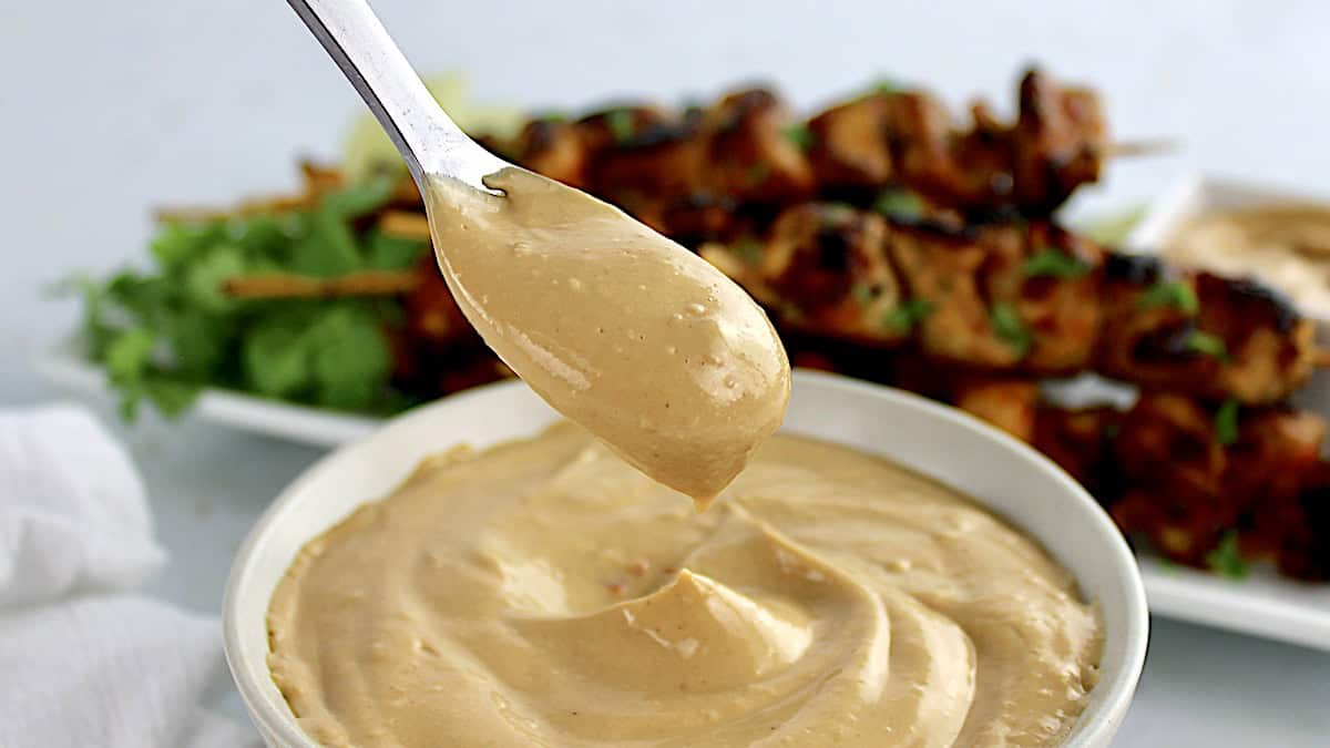 Peanut Sauce being spooned out of white bowl with chicken skewers in back