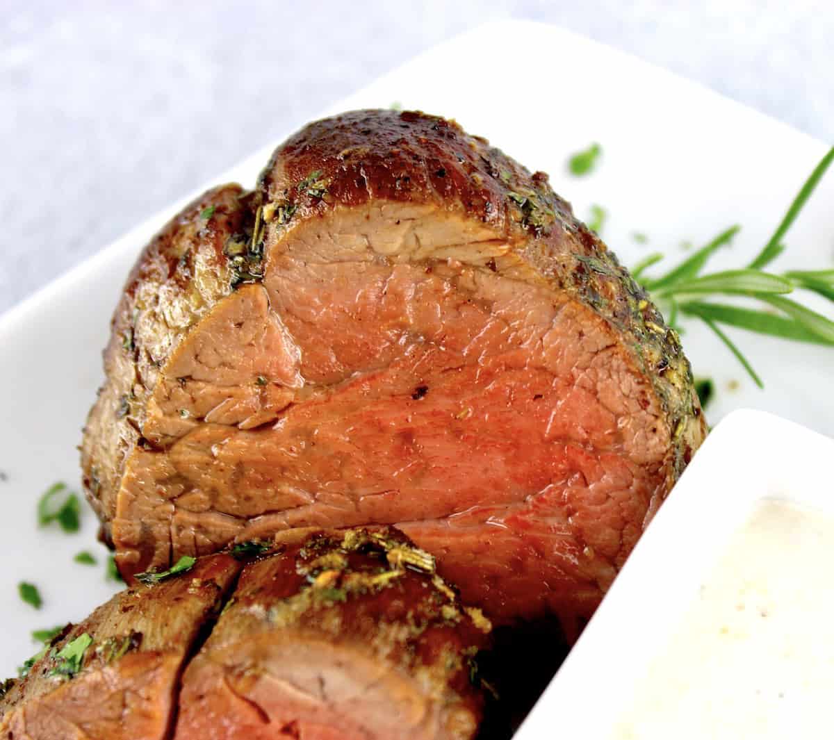 closeup of slice of Roast Beef Tenderloin on white plate with rosemary in background