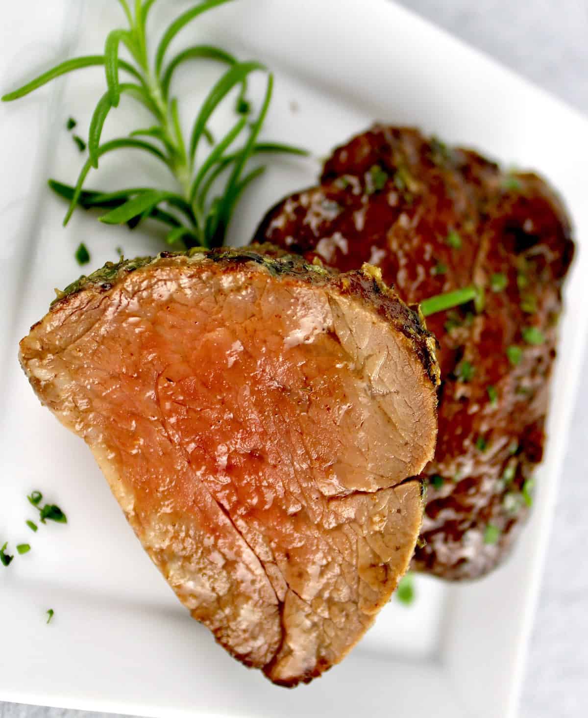 2 Roast Beef Tenderloin slices one with cut side up on white plate with rosemary sprig