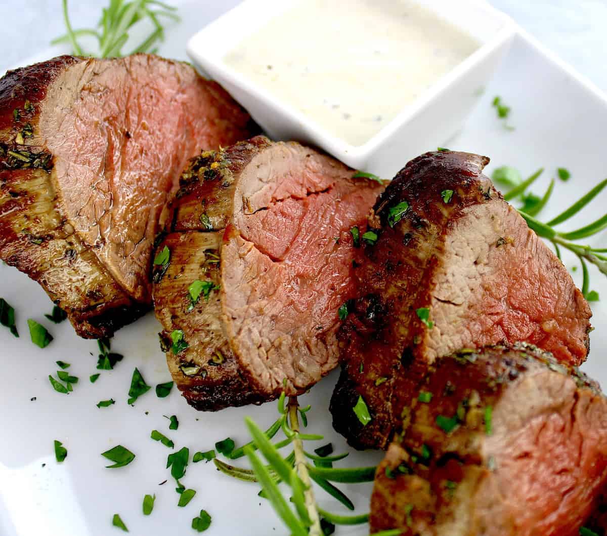 Roast Beef Tenderloin slices on white plate with fresh rosemary and creamy horseradish sauce in white square bowl