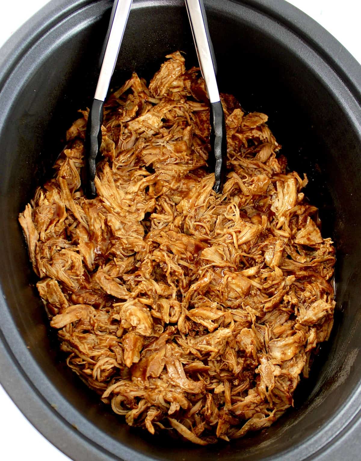 pulled pork in slow cooker insert with black tongs