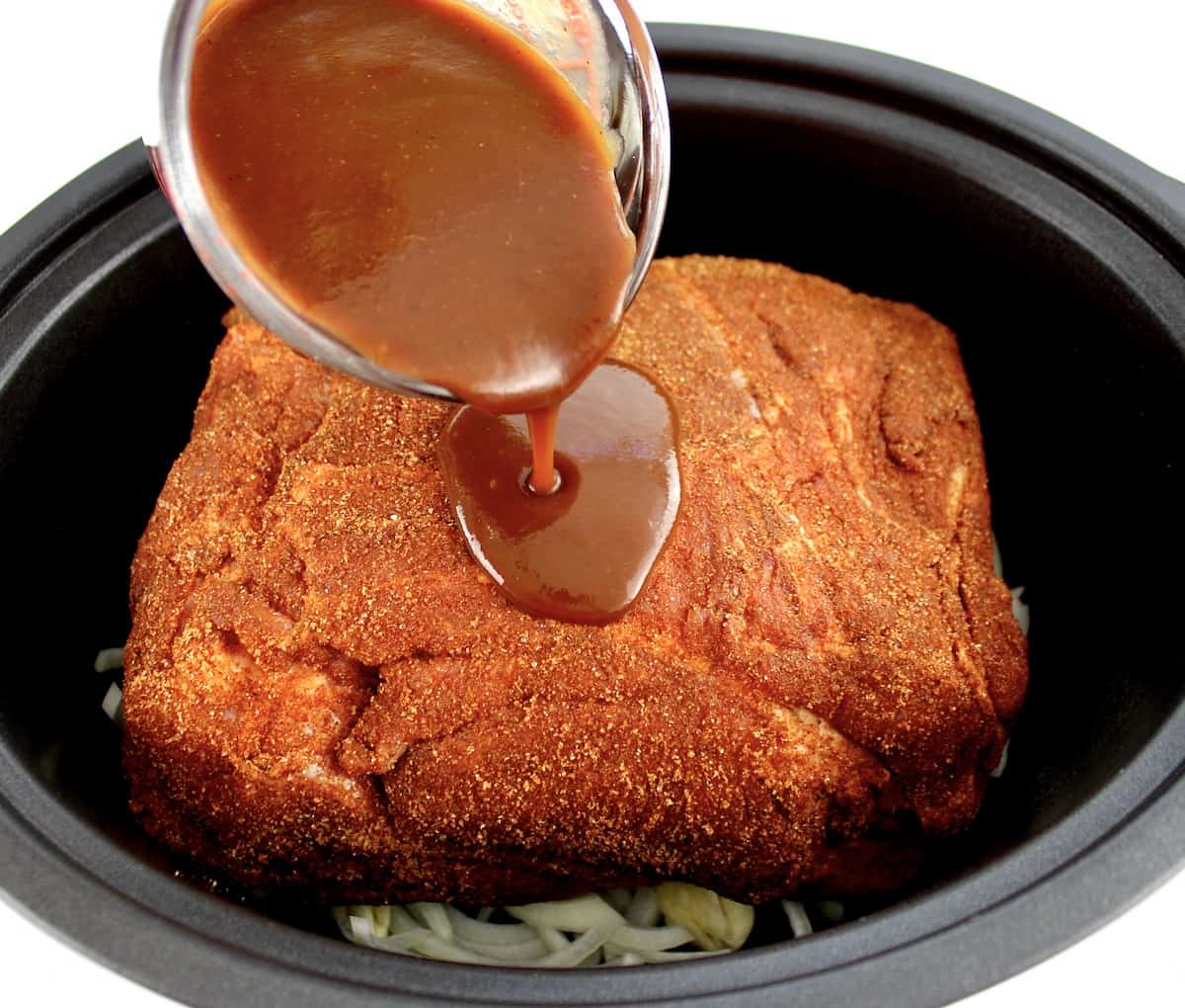 raw pork shoulder with bbq spice rub on it in slow cooker insert with bbq sauce being poured on top