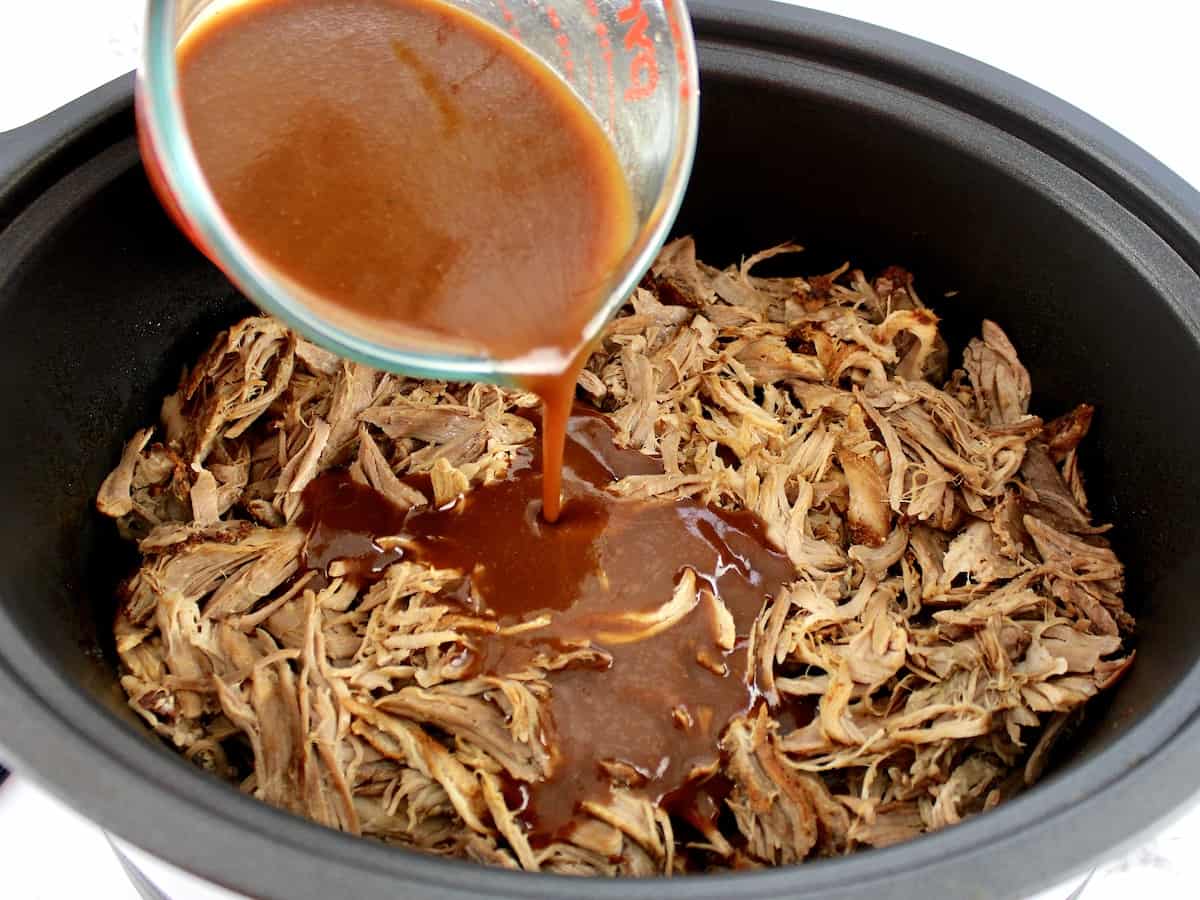 pulled pork in a slow cooker with bbq sauce being poured over