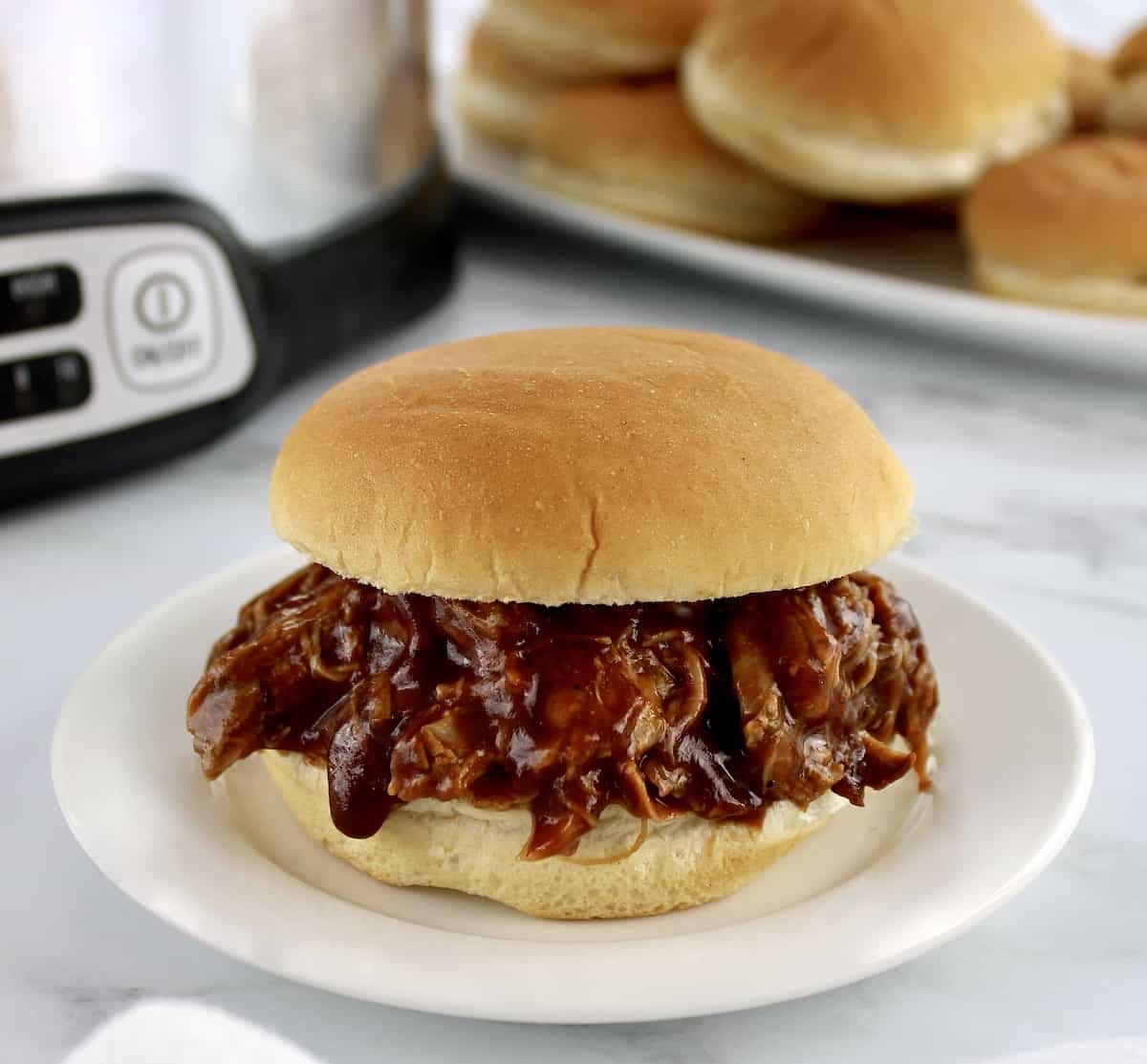 Slow Cooker Pulled Pork sandwich on white plate with buns and slow cooker in background