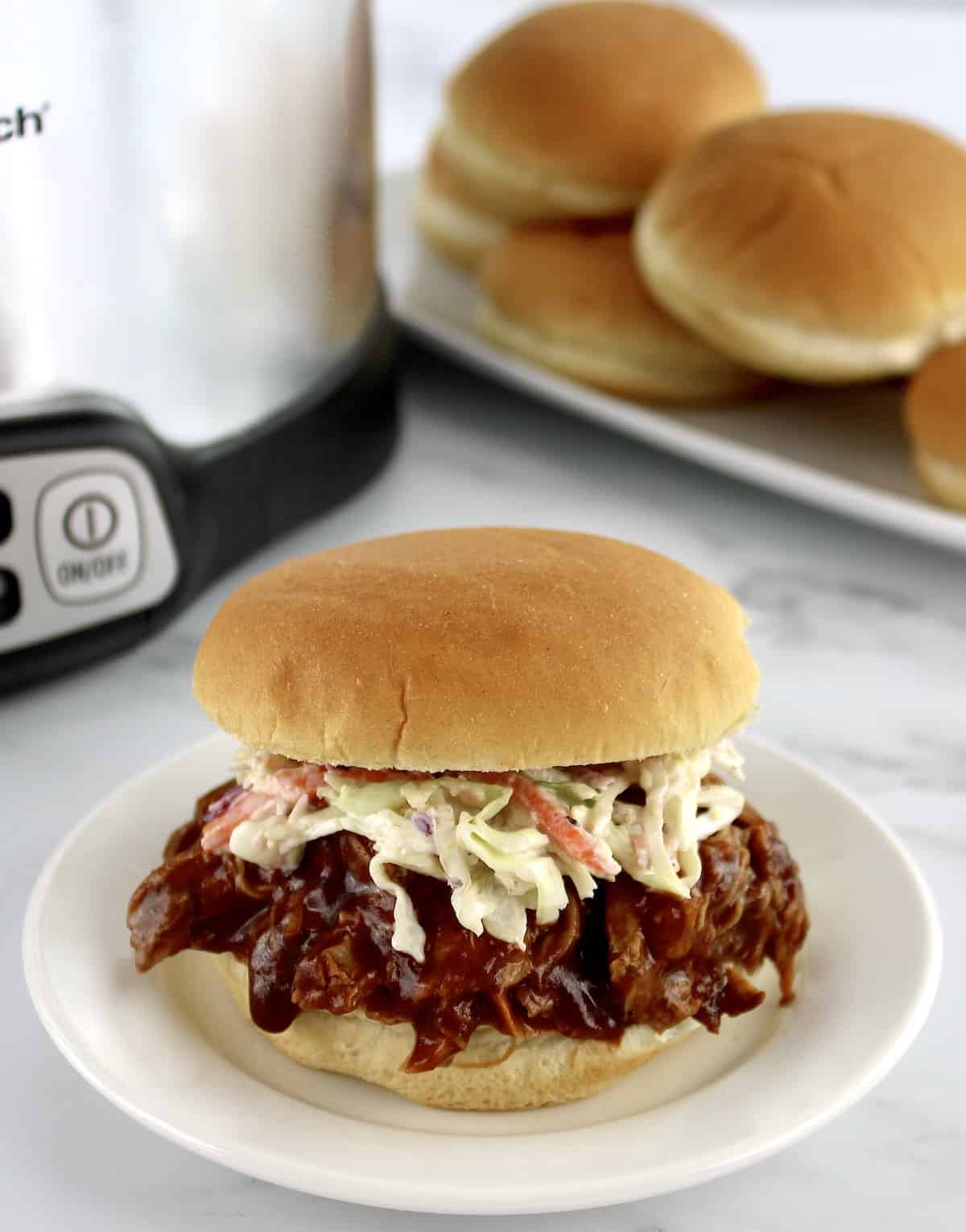 pulled pork sandwich with coleslaw on white plate with buns and slow cooker in background