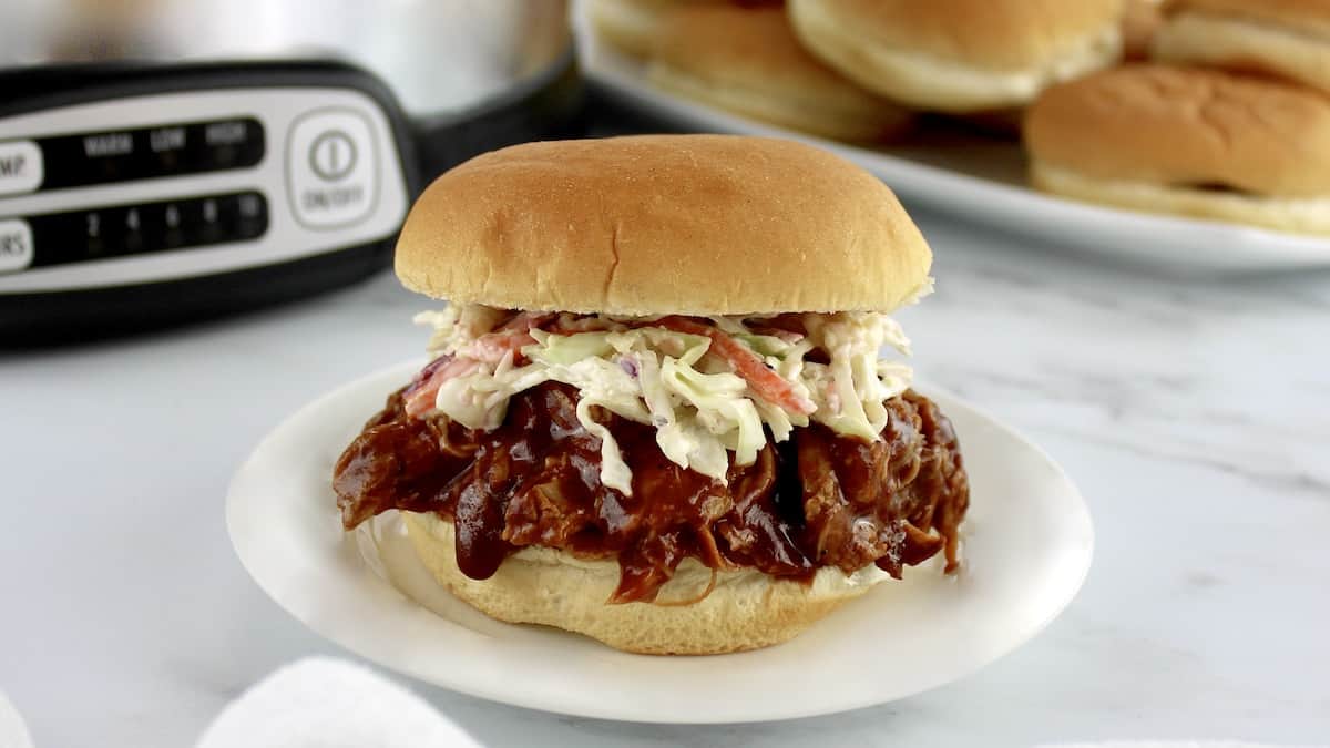 pulled pork sandwich with coleslaw on white plate with buns and slow cooker in background