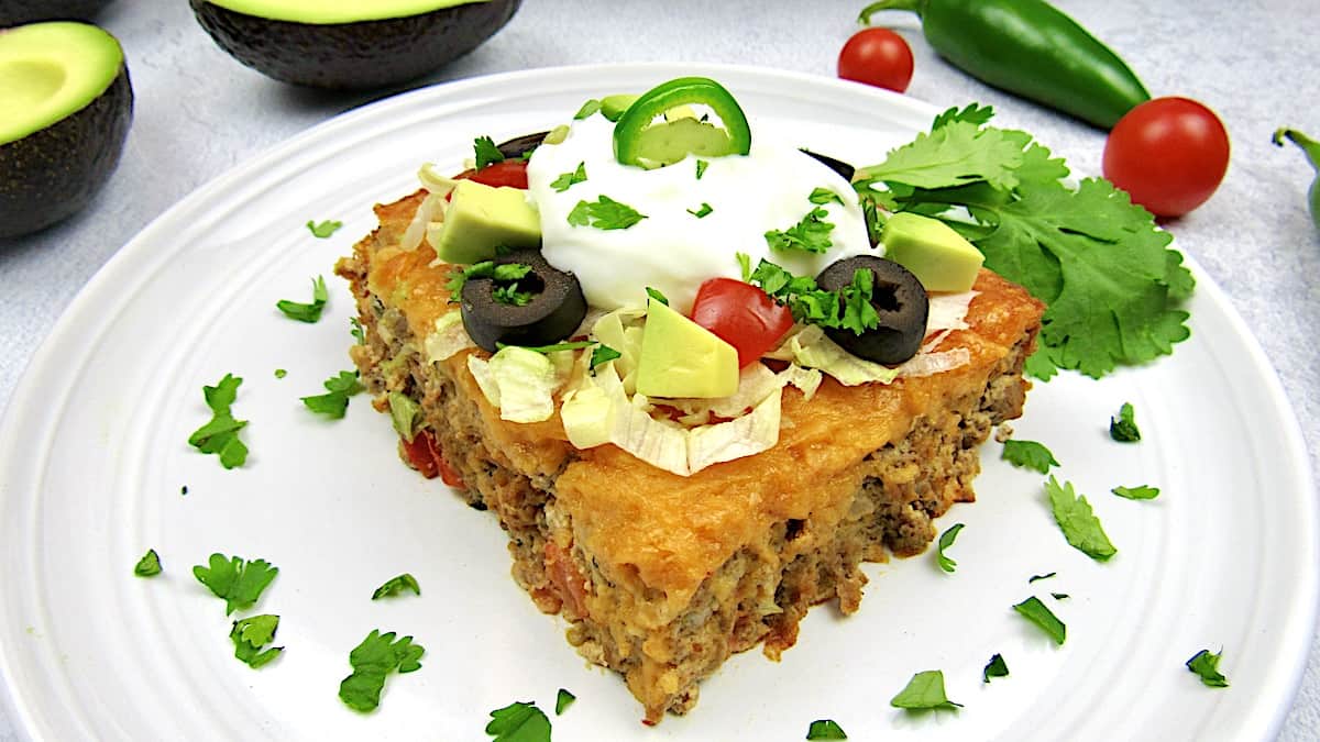 Taco Casserole slice on white plate with olives, diced avocado and sour cream on top