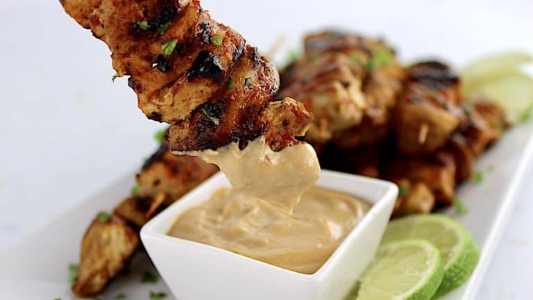 thai chicken satay dipping into sauce on white plate with lime slices on side