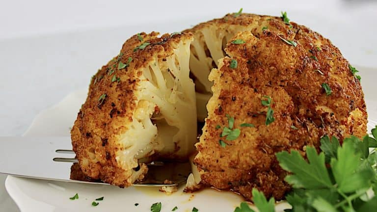 Whole Roasted Cauliflower on white plate with chopped parsley and piece being pulled out