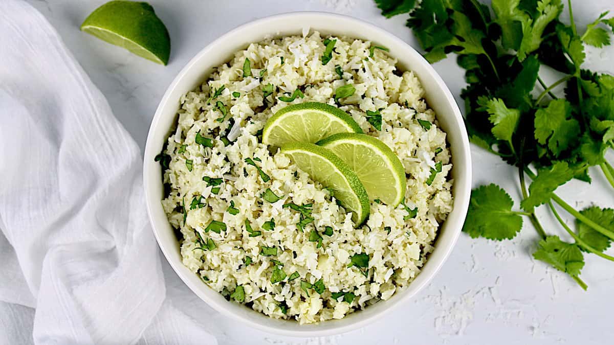 closeup of coconut cauliflower rice in white bowl with 3 lime slices on top and cilantro in back