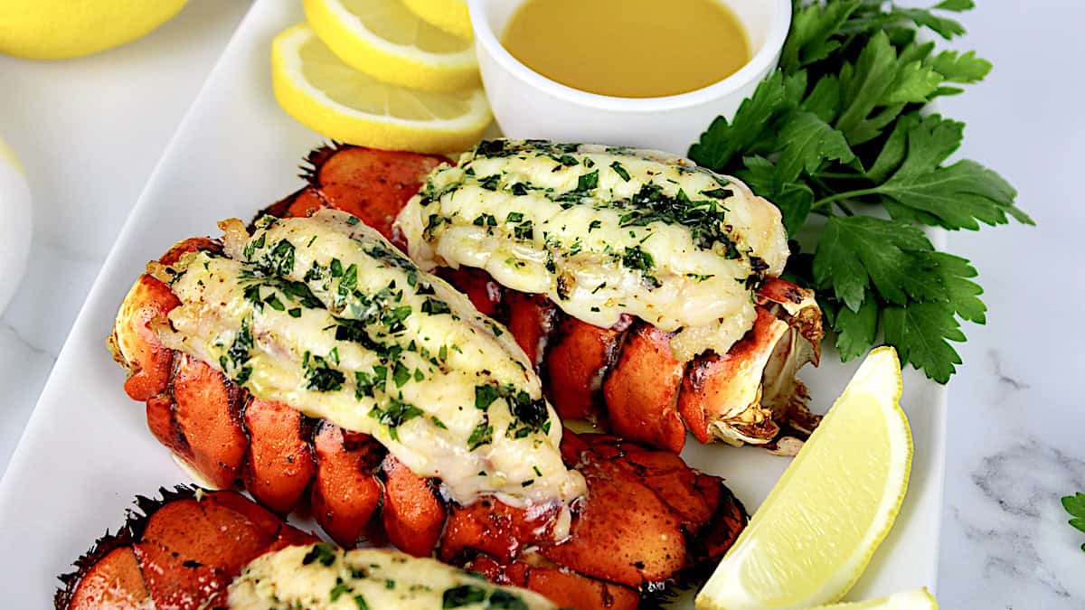 2 Broiled Lobster Tails on white plate with melted butter in cup with lemon slices