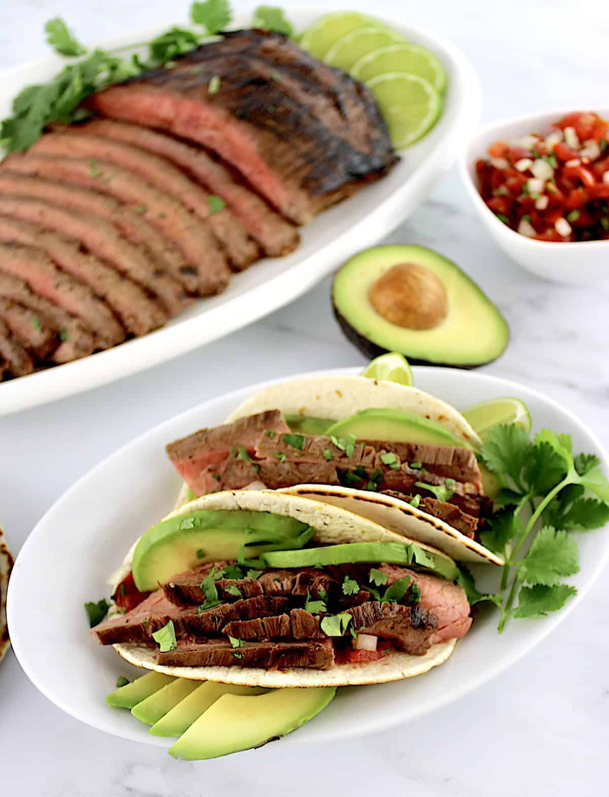 Carne Asada in 2 tacoes with avocado slices with sliced flank steak in background