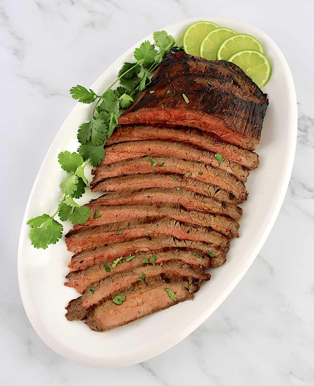 Carne Asada sliced on white oval platter with fresh cilantro with 4 lime slices