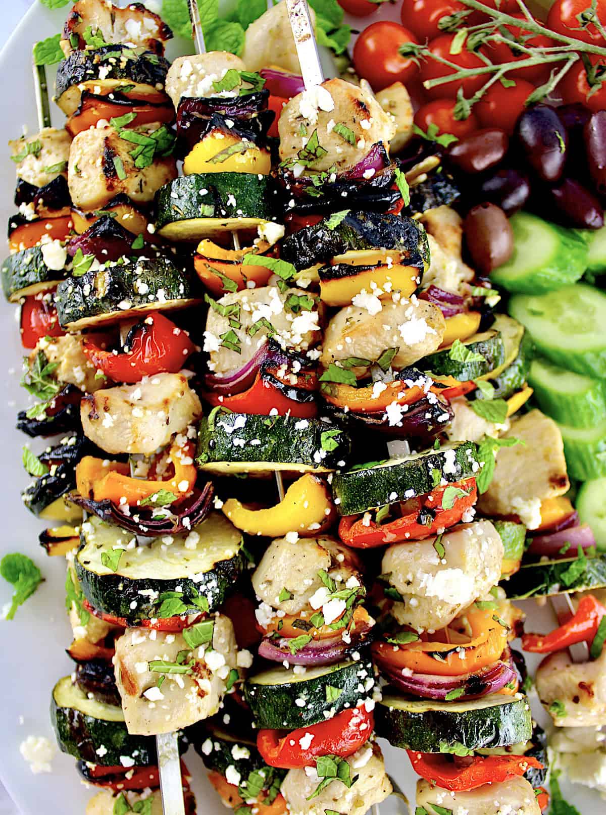 closeup of Greek Chicken Kebabs with cucumbers, tomatoes and olives in background