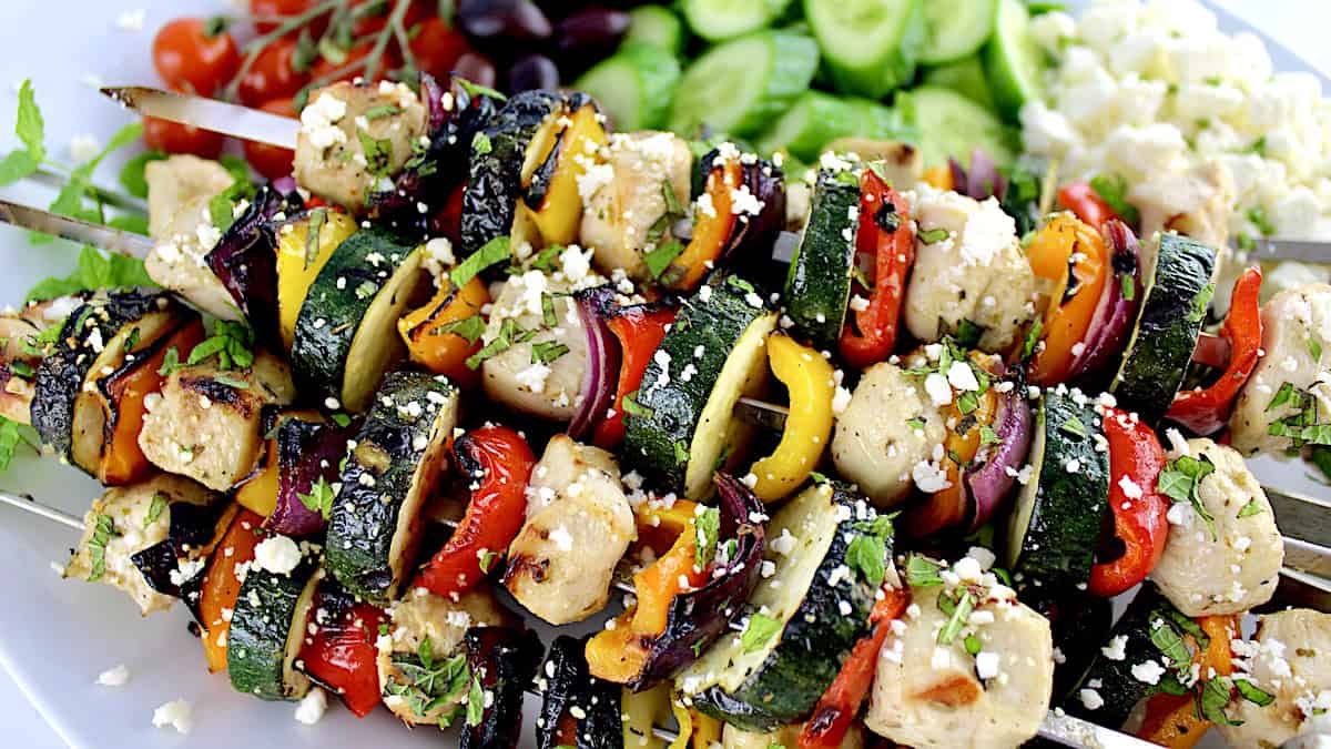 Greek Chicken Kebabs on white plate with sliced cucumbers, olives and tomatoes in background