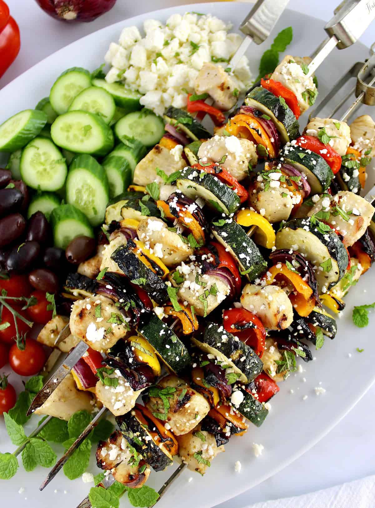 closeup of Greek Chicken Kebabs with cucumbers, tomatoes and olives in background