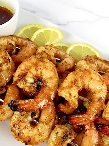closeup of Grilled Cajun Shrimp Skewers on white platter with dipping sauce in cup and lemon slices