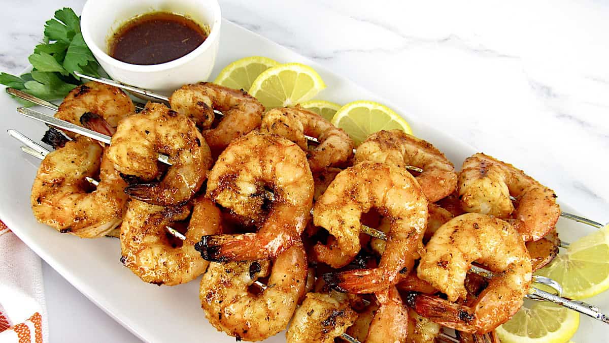 closeup of Grilled Cajun Shrimp Skewers on white platter with dipping sauce in cup and lemon slices