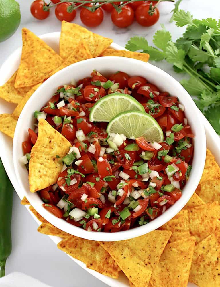 Pico de Gallo in white bowl with tortilla chip and 2 lime slices with more chips around the bowl