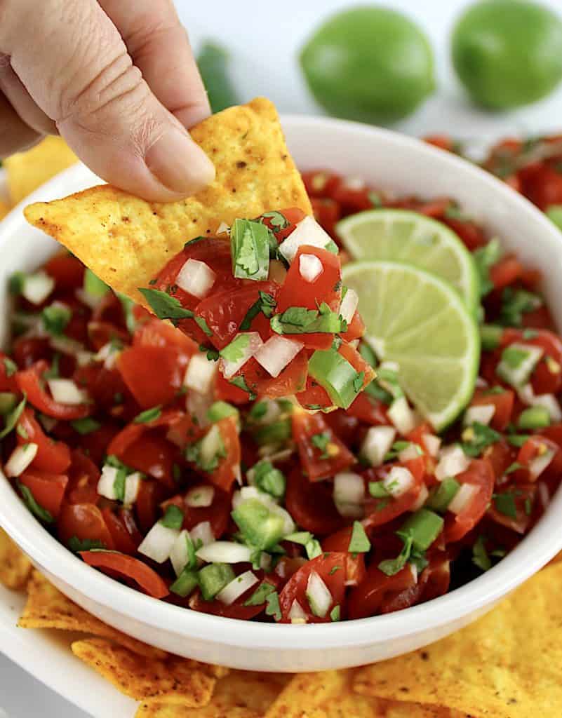Pico de Gallo in white bowl with 2 lime slices with tortilla chip being scooped up