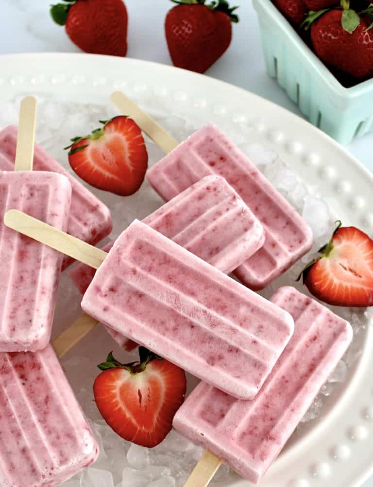 Strawberry Coconut Cream Popsicles on white platter on crushed ice and halved strawberries