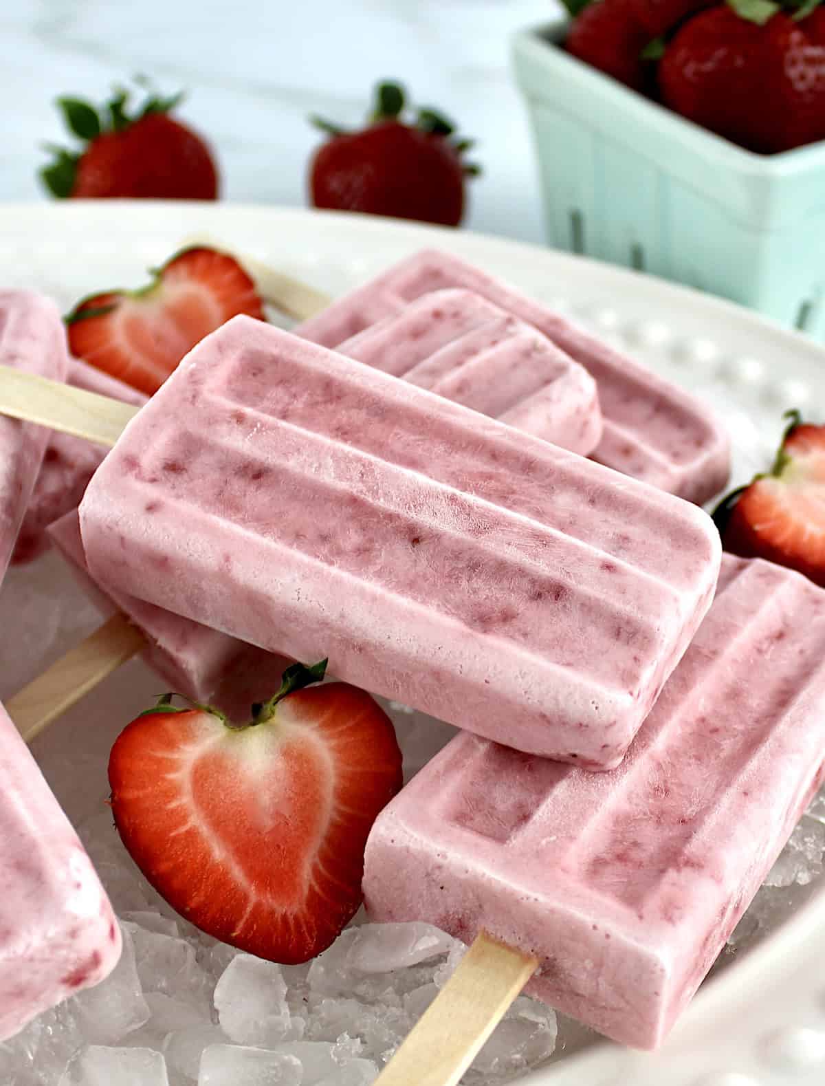 Strawberry Coconut Cream Popsicles on white platter on crushed ice and halved strawberries