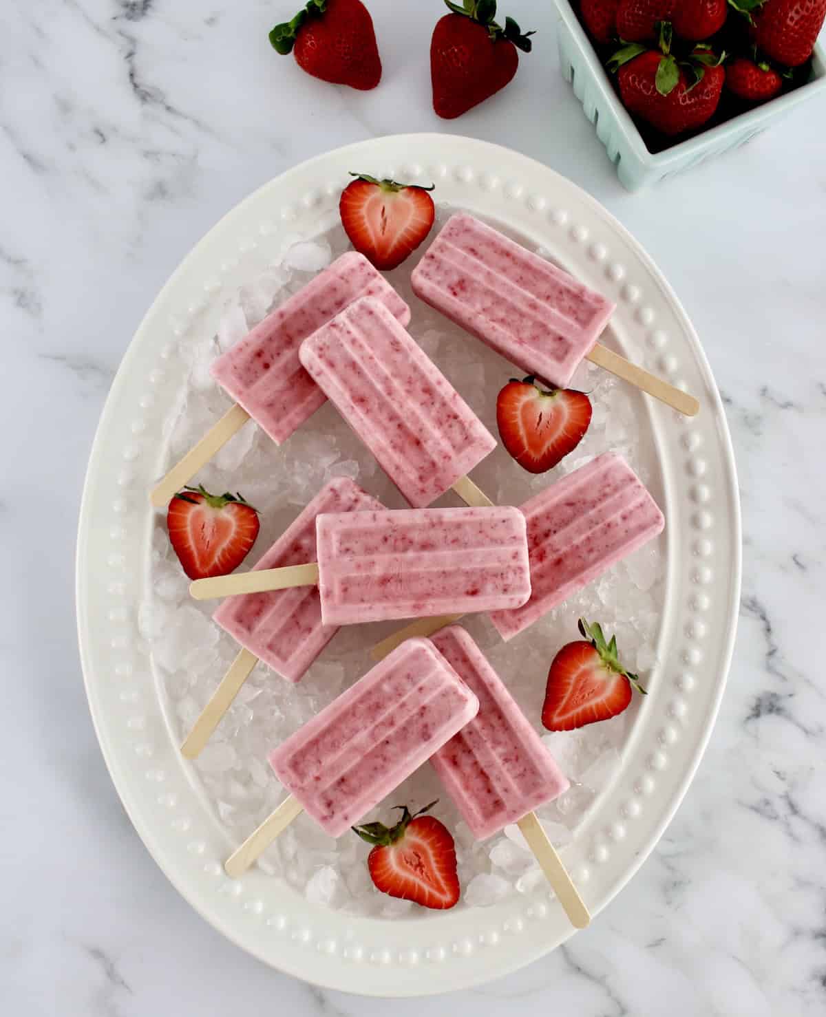overhead view of Strawberry Coconut Cream Popsicles on white oval platter on crushed ice and strawberry halves
