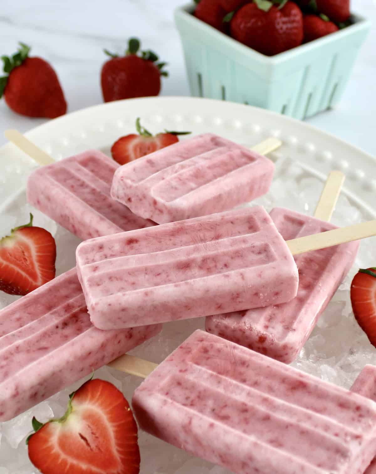 Strawberry Coconut Cream Popsicles on white platter on crushed ice and halved strawberries
