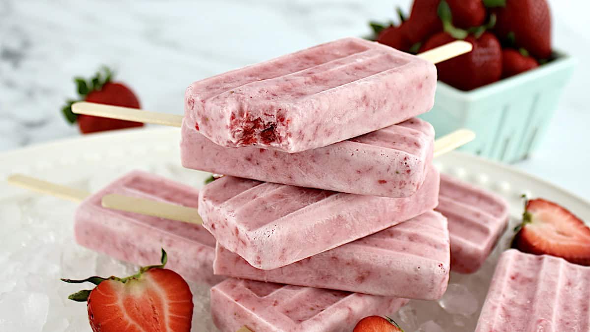 Strawberry Coconut Cream Popsicles stacked up on white platter with bite missing from top popsicle