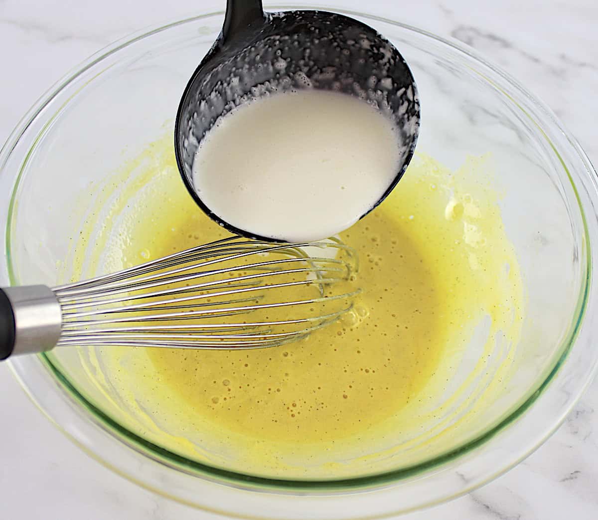 cream being ladled into glass bowl with egg yolks sweetener and whisk