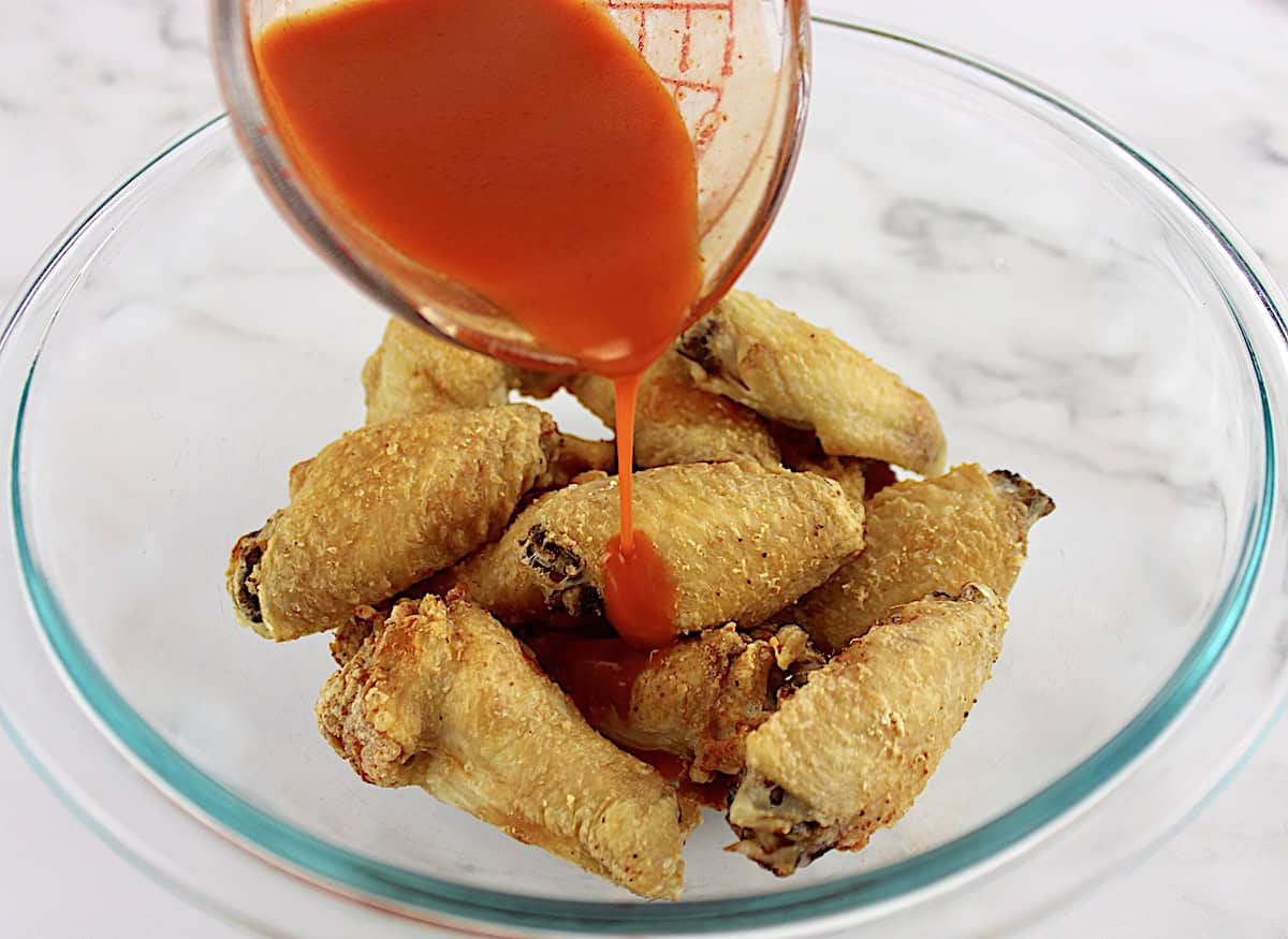 crispy chicken wings in glass bowl with wing sauce being poured over top