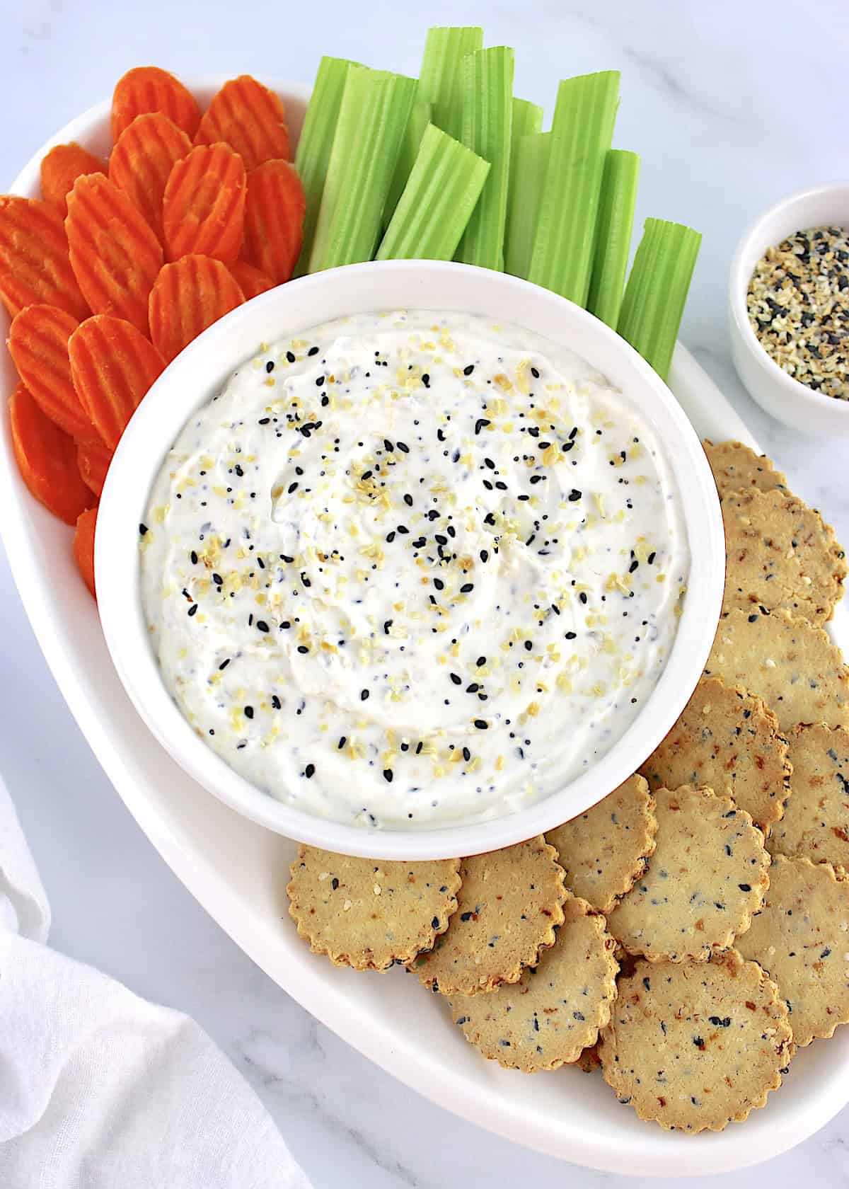 Everything Bagel Dip in white bowl with crackers, celery sticks and carrot chips on white oval platter