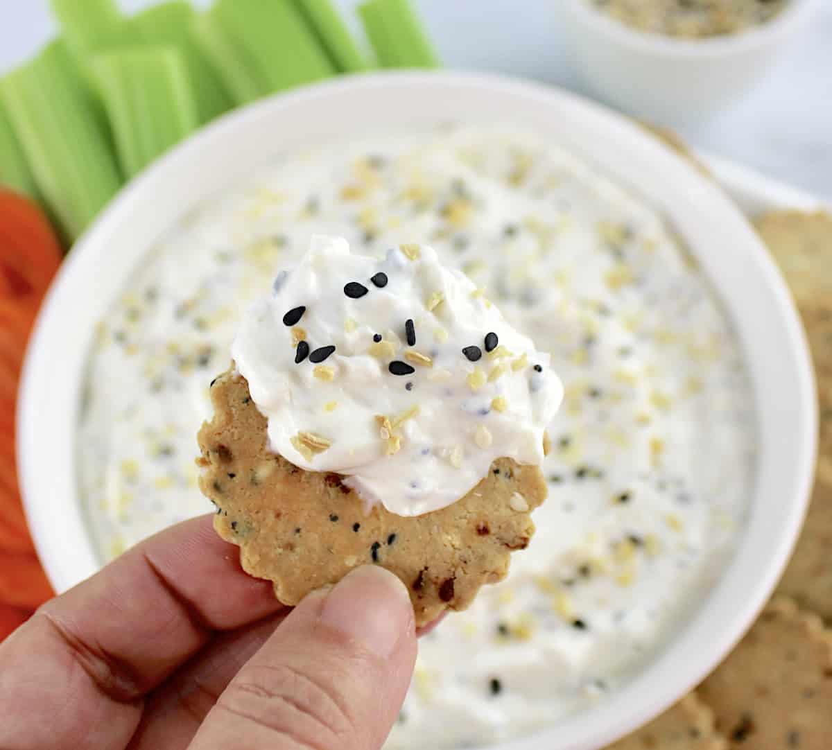 Everything Bagel Dip in white bowl with cracker dipped in and held up