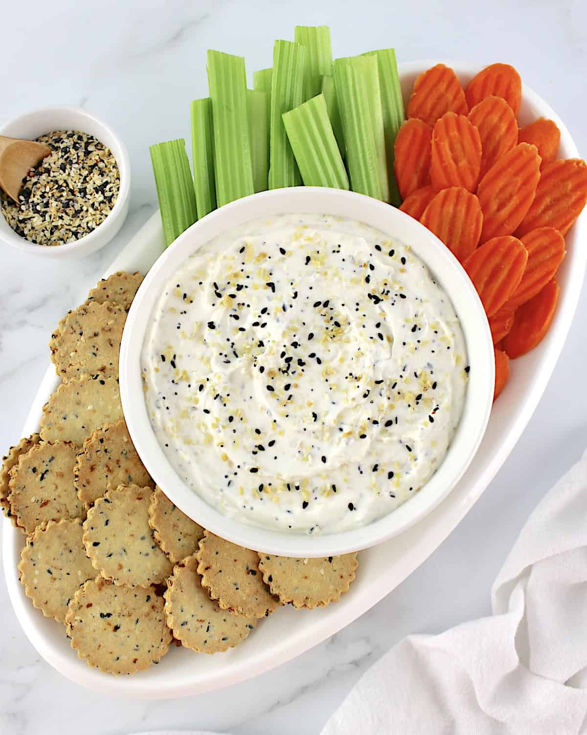 Everything Bagel Dip in white bowl with crackers, celery sticks and carrot chips on white oval platter