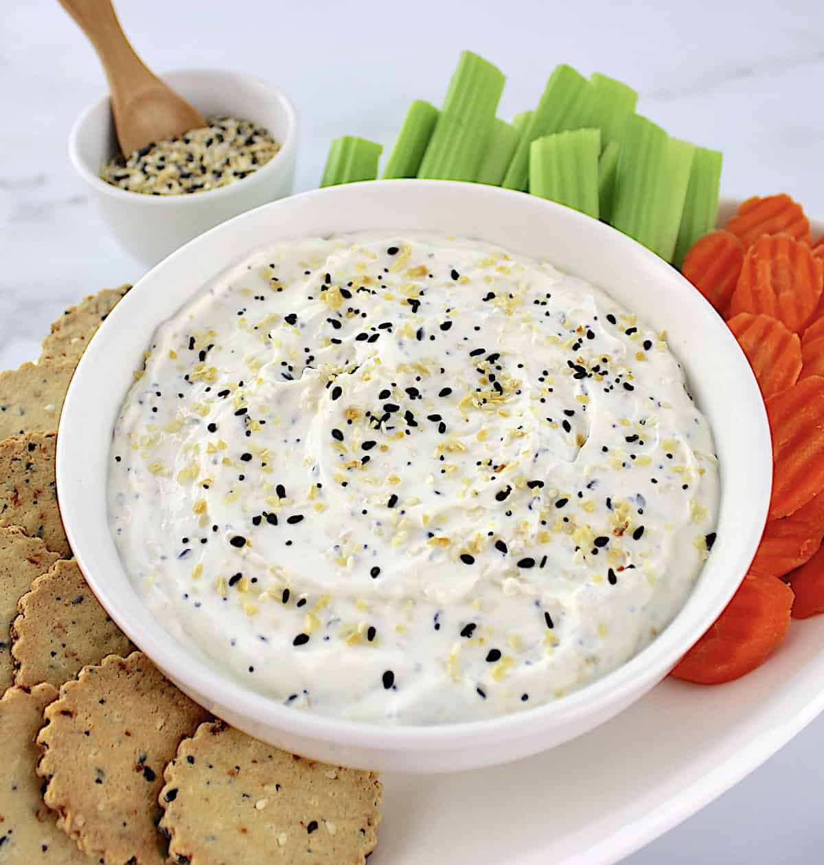 Everything Bagel Dip in white bowl with crackers, celery sticks and carrot chips on white oval platter