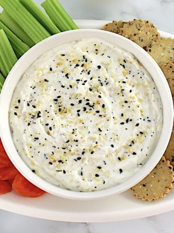 Everything Bagel Dip in white bowl with crackers, celery sticks and carrot chips on white oval platter