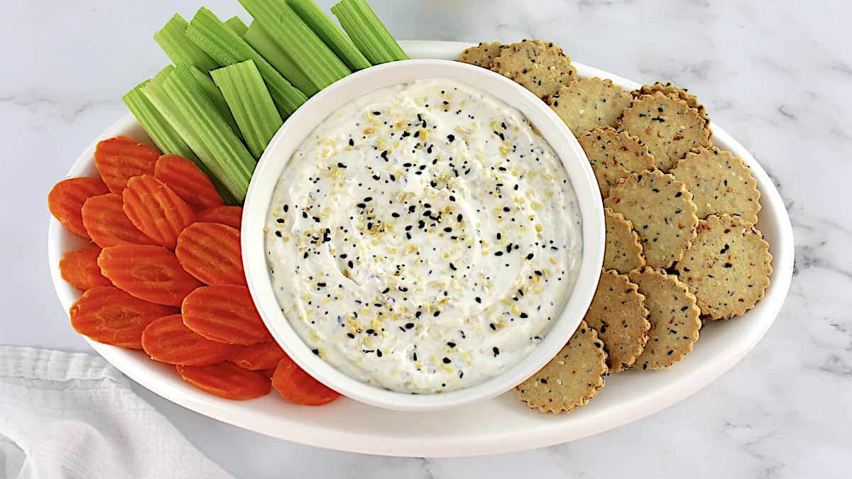 Everything Bagel Dip in white bowl with crackers, celery sticks and carrot chips on white oval platter