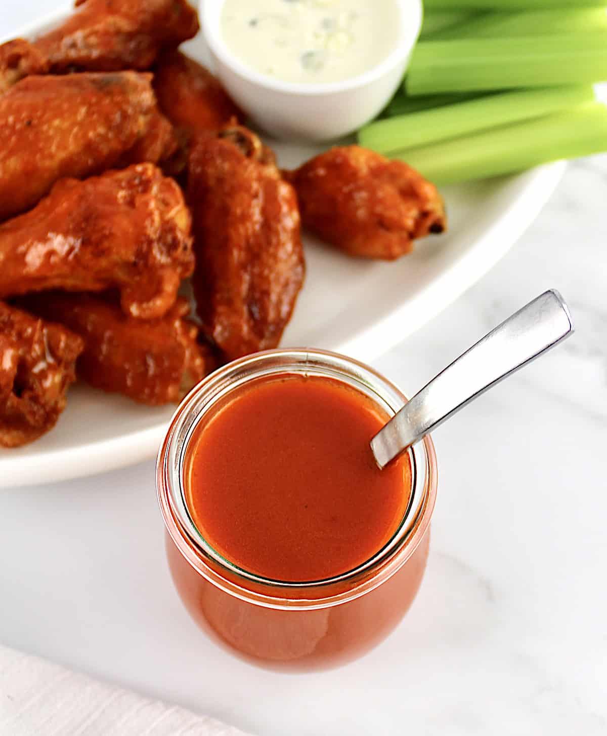Homemade Buffalo Wing Sauce in open glass jar with spoon and wings in back