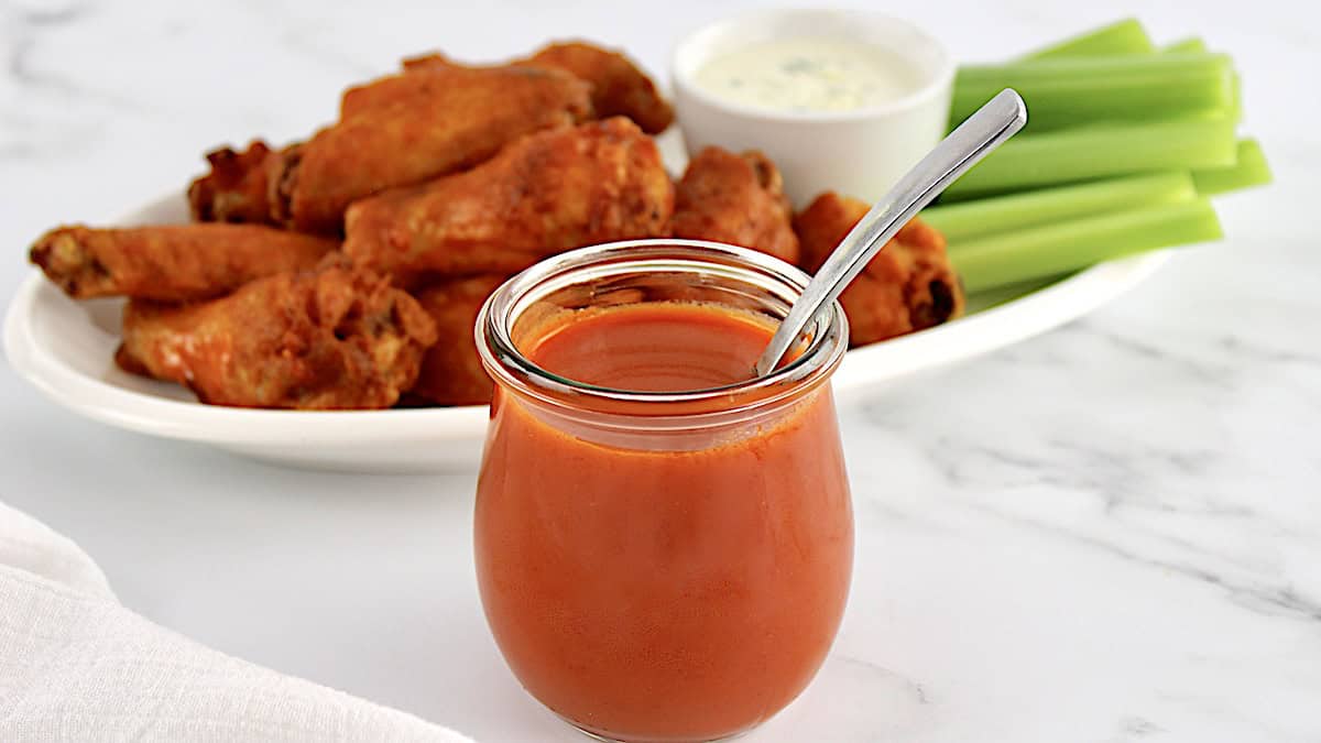 Homemade Buffalo Wing Sauce in open glass jar with spoon and wings in back