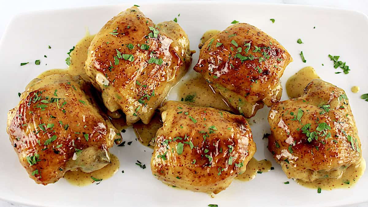 overhead view of Honey Mustard Baked Chicken Thighs on white plate with sauce and chopped parsley on top