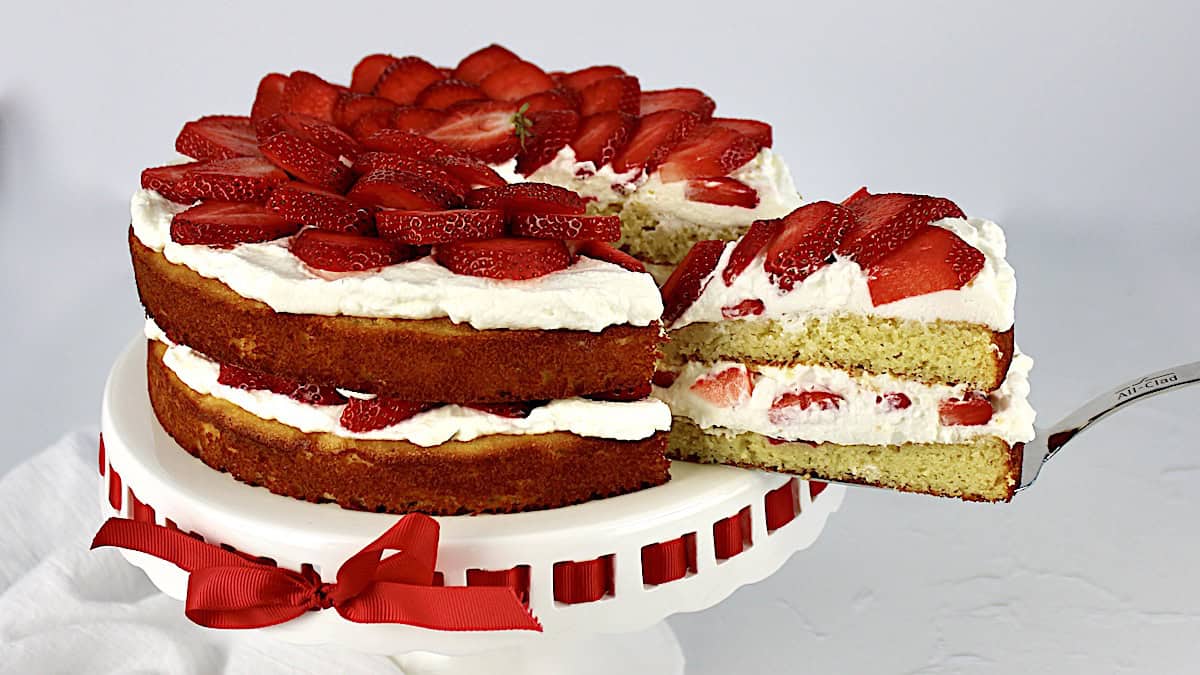 Keto Strawberry Shortcake on white cake stand with slice being removed using silver cake server