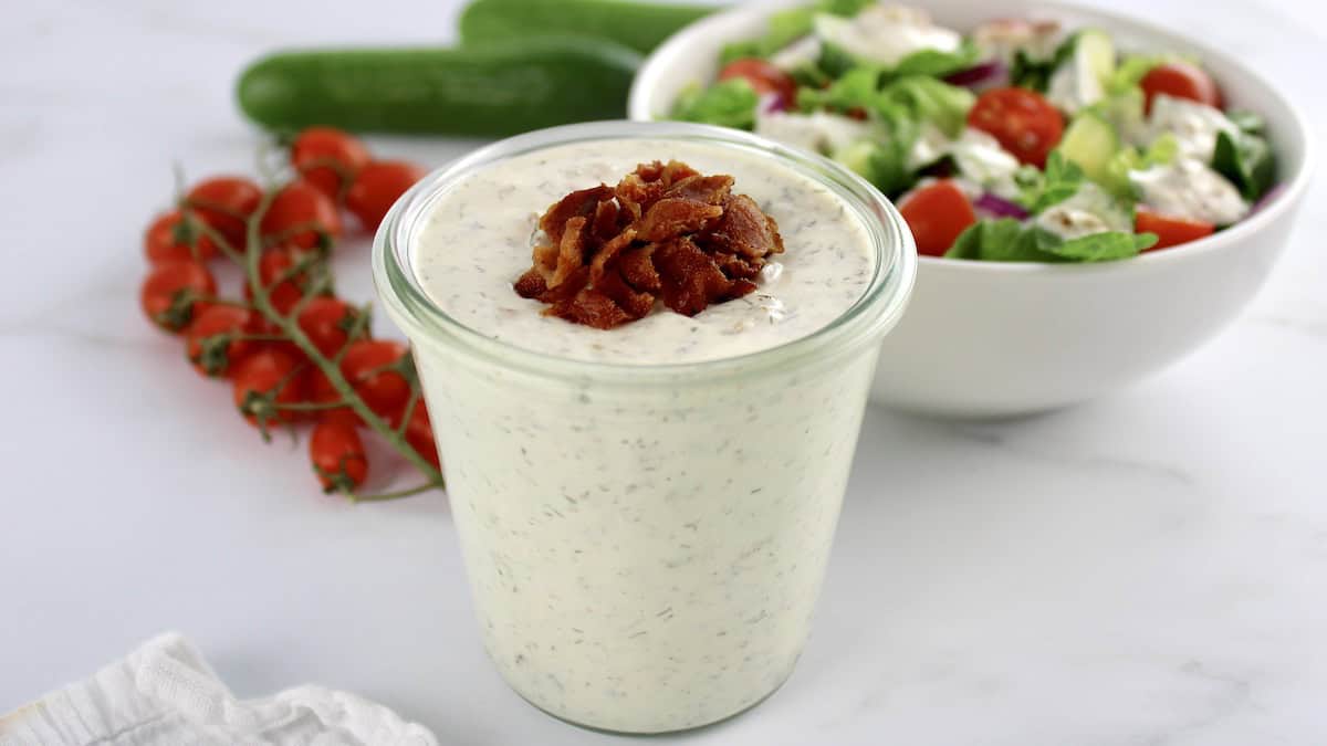 Bacon Ranch Dressing in open glass jar with crumbled bacon on top and salad in back