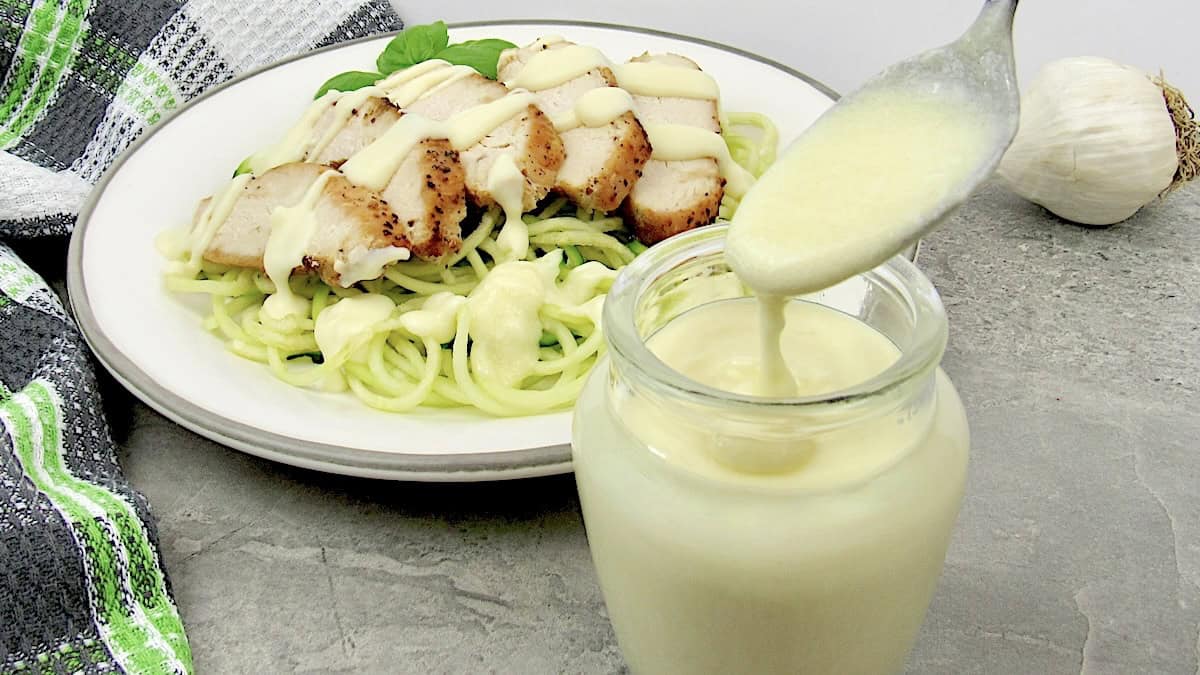 Easy Keto Alfredo Sauce being spooned out of glass jar with sliced chicken over zucchini noodles with sauce over in back