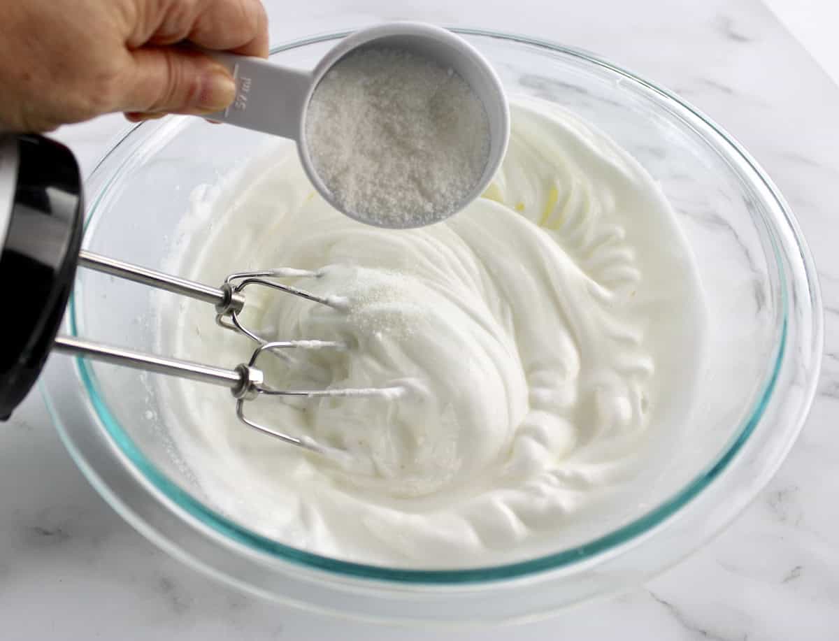 whipped egg whites in glass bowl with hand mixer and sweetener being poured in slowly