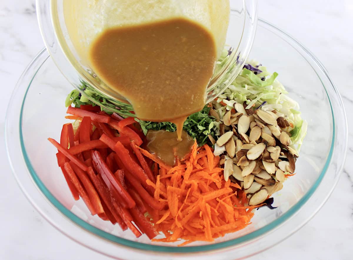 Peanut Ginger Slaw ingredients in groups in glass bowl umixed with dressing being poured over top